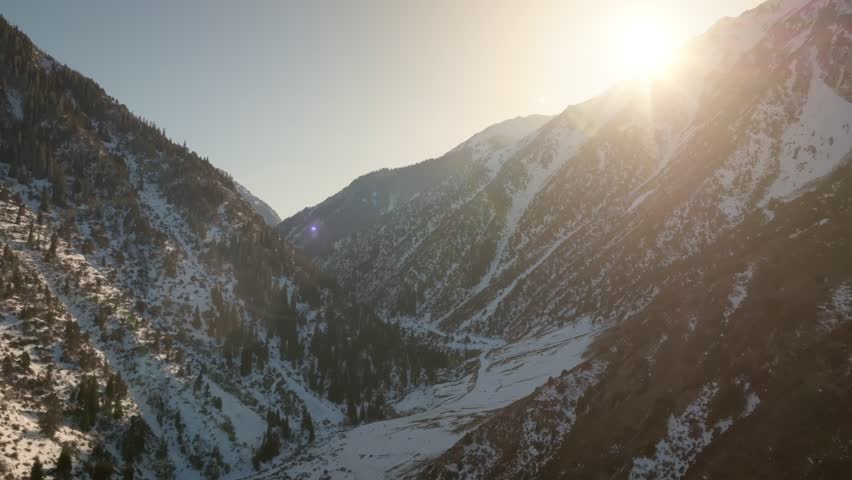 Aerial footage of sunrise light and lens flare over snow covered mountain slopes and forested valley in Kegety Gorge, Kyrgyzstan, scenic winter nature
