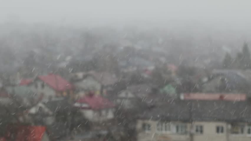 Aerial view of a small town with red rooftops covered in falling snow during a heavy snowfall, foggy and gray winter sky above.