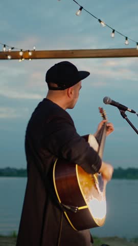 Man standing with acoustic guitar under string lights by a lakeside at dusk