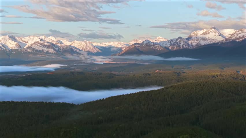 Sideways panning aerial drone footage of the Canadian Rockies near Banff National Park with morning fog and a temperature inversion over the forested mountains.