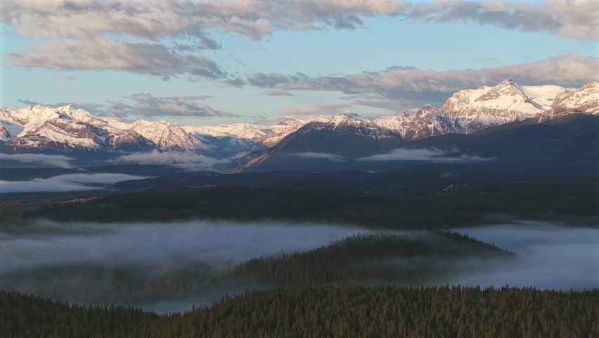Climbing and panning aerial drone footage of the Canadian Rockies near Banff National Park with a morning temperature inversion and fog over the forest.