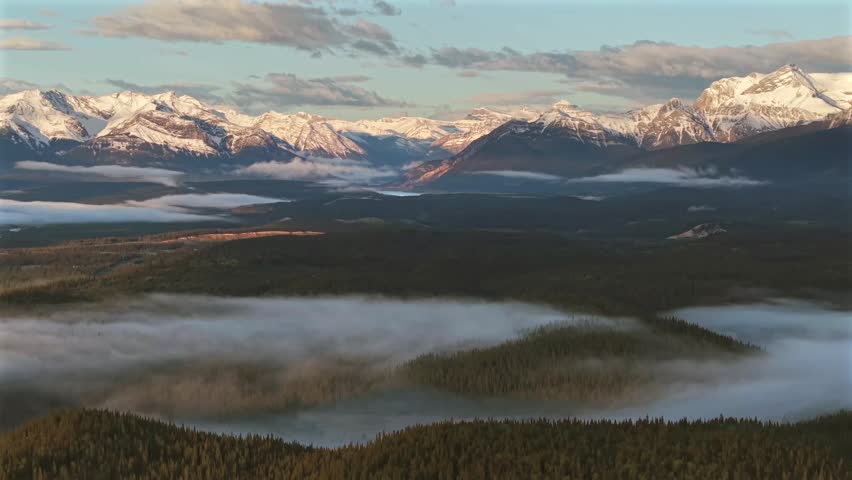 Descending aerial drone footage of the Canadian Rockies near Banff National Park with morning fog and a temperature inversion over pine forest.