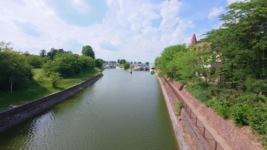 Siofok Hungary May 31 2026. Elevated view of a canal waterway bordered by trees and stone embankments leading toward Lake Balaton on a sunny day. Scenic urban waterfront footage featuring water management infrastructure and green surroundings.