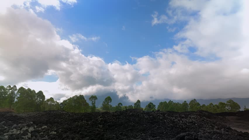 Timelapse of moving white clouds over Mount Batur black lava fields and green trees in Kintamani, Bali