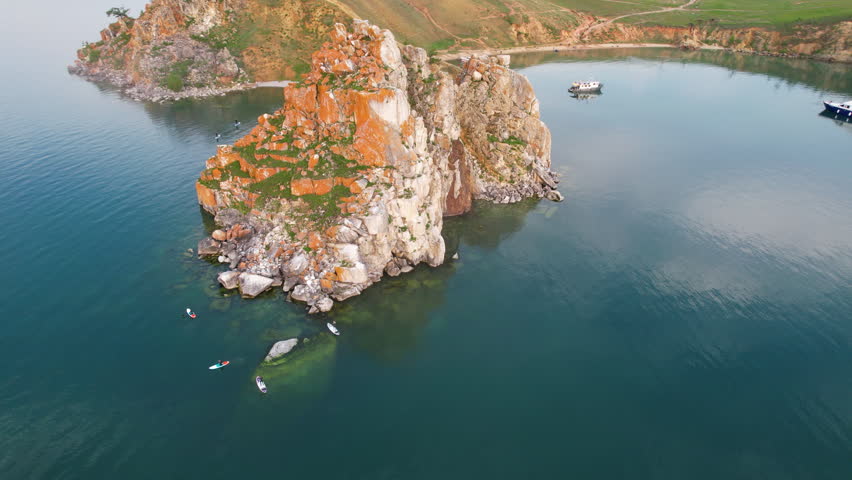 Lake Baikal on a summer evening. Flight over Cape Burkhan or Shamanka Rock on Olkhon Island. Tourists paddleboard in the bay. Active recreation. 