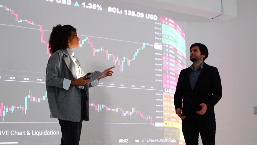 A businessman and a Latina female investment advisor stand speaking in front of a room of stock and investment analysis experts in a company seminar room.
