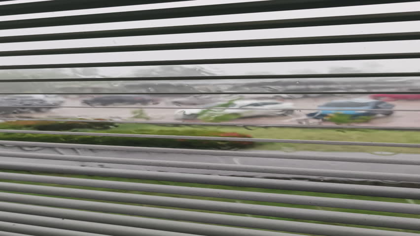 A view through window blinds shows a parking lot during a heavy rainstorm outdoors