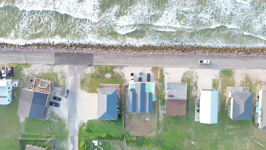 Seawall coastal row Surfside Beach homes along Seashell Dr line rock barrier beside Gulf corridor with elevated structures facing open water. Sunlit roofs, bright surf glow in Freeport, Texas. USA