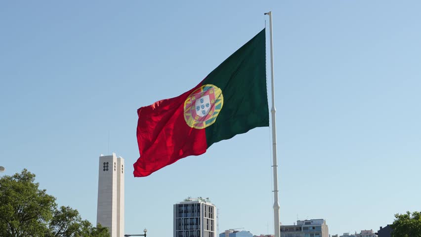 The big national flag of Portugal waving on a tall flagpole against a blue sky, with modern buildings and monument columns visible below near Edward VII Park in Lisbon. Wide shot