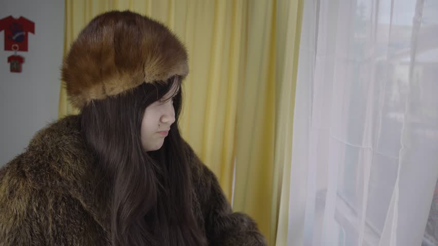 Close up of young woman in winter fur coat and hat looking through window curtains