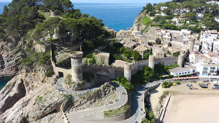 Aerial view of Tossa de Mar, a historic Costa Brava town crowned by its medieval walls, overlooking a beautiful sandy beach and turquoise Mediterranean waters. 