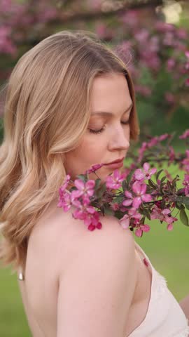Soft close-up of a young blonde woman with closed eyes near pink flowers in a blurred outdoor garden, lit by gentle natural daylight with a calm and serene mood