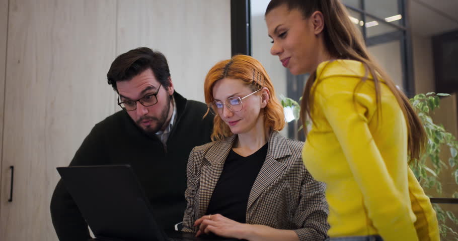 Three colleagues gather in an office to discuss a project. They focus on a laptop screen while sharing ideas and looking engaged. The atmosphere shows teamwork and collaboration.
