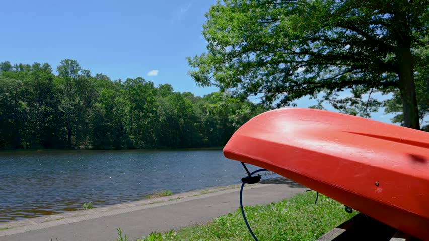 Red kayak canoe on rack by the shore of a flowing Pennsylvania lake lush green foliage. Idyllic woodland background calm flowing water blue sky. Beautiful scenic state park with copy space. Vacation travel tourism visit PA landscape nature background. High quality 4k footage