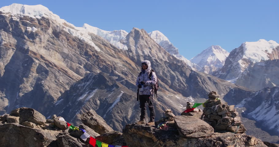 Gokyo Ri Nepal, orbit drone shot of a lone trekker standing among prayer flags on the rocky summit viewpoint looking toward Cholatse peak, surrounded by Everest Region peaks