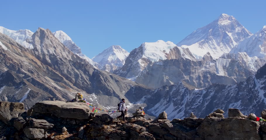 Gokyo Ri, Everest Nepal, orbit drone shot of a trekker walking along the rocky path towards the mountain summit viewpoint, surrounded by prayer flags and Himalayan peaks