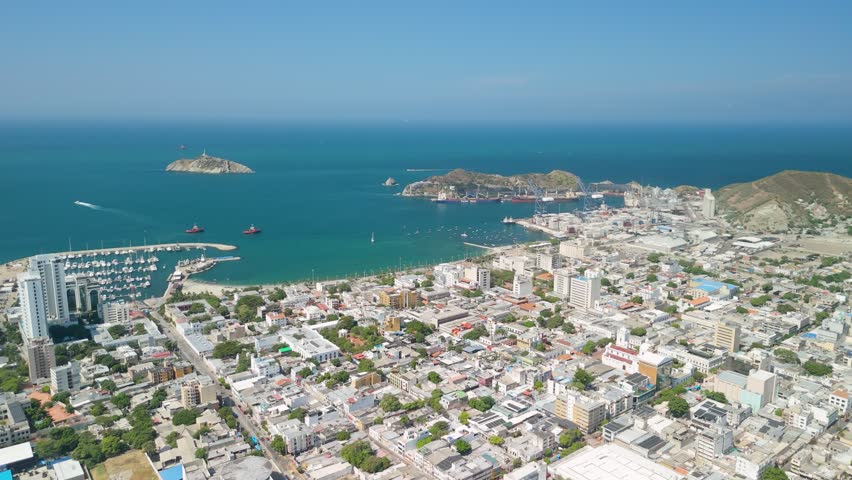 High-altitude aerial flyover over Santa Marta city revealing marina, Bahía de Santa Marta, Isla El Morro lighthouse island and industrial port