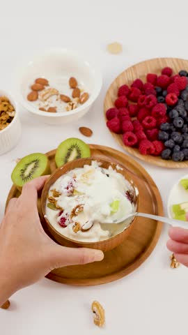 Overhead shot of hands scooping yogurt with fruit and nuts from a wooden bowl.