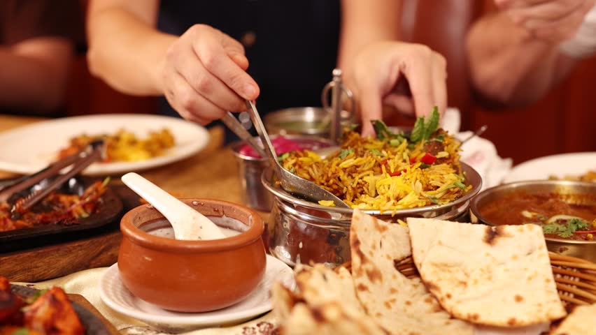 Close-up of hands serving yellow biryani rice from metal bowl alongside naan and curry.