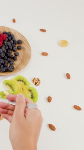 Overhead shot of hand scooping yogurt with berries, kiwi, and nuts from wooden bowl.