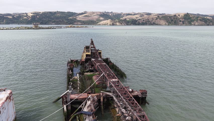Cinematic aerial drone footage flying low along the length of an abandoned, rusting crane barge wreck off the Benicia waterfront, California, the collapsing steel structure leading the eye out across the Carquinez Strait toward the hills and the Carquinez Bridge. A moody, atmospheric study of maritime decay in tidal water.