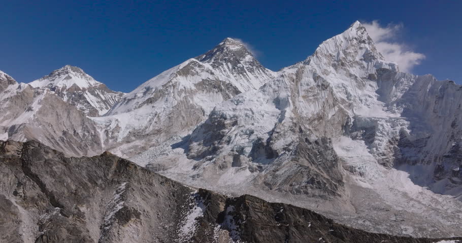 Kala Patthar Everest BaseCamp Nepal, drone gliding shot of towering snow-capped Himalayan giants featuring Mount Everest and Nuptse rising above dark rocky ridges under a clear blue sky.