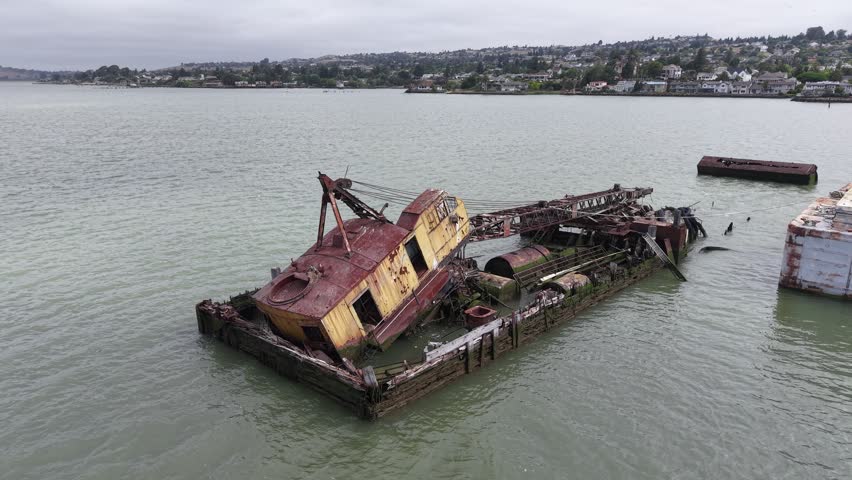 Cinematic aerial drone footage orbiting an abandoned, rusting crane barge wreck and a derelict grey barge off the Benicia waterfront, California. Decaying steel and timber sit in the shallow Carquinez Strait, with the waterfront homes, palms, and rolling Northern California hills rising behind under an overcast sky.