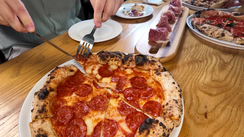 Close-up of hands slicing a wood-fired pepperoni pizza on a wooden table with charcuterie.