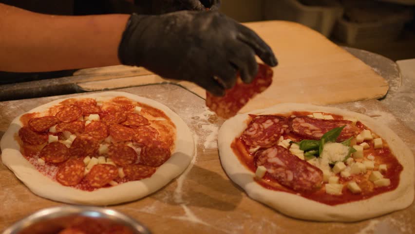 Chef in black gloves adds pepperoni and olive oil to raw pizza dough in kitchen.