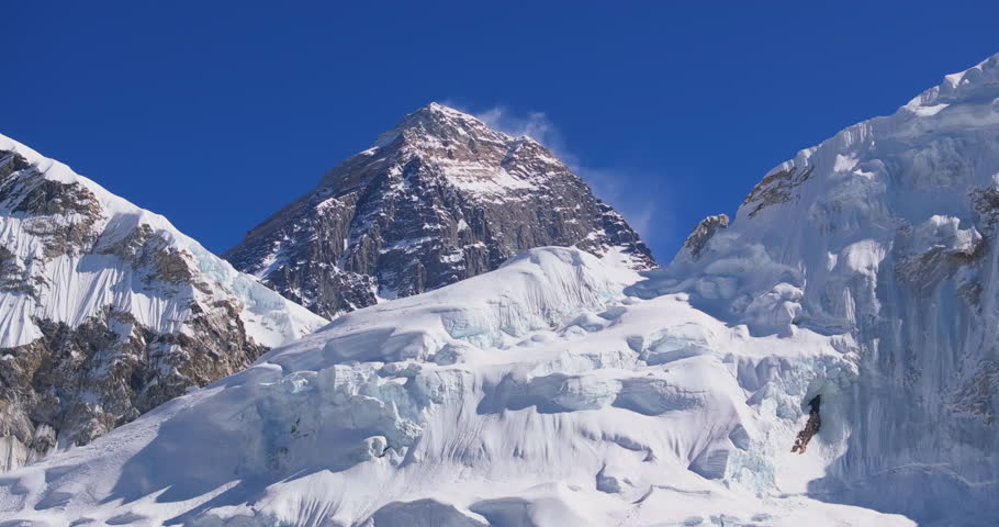 Everest region, Nepal, drone aerial sideways glide shot capturing the dark rocky peak of Mount Everest with a blowing snow plume at the summit rising behind the snow-capped slopes of Nuptse.