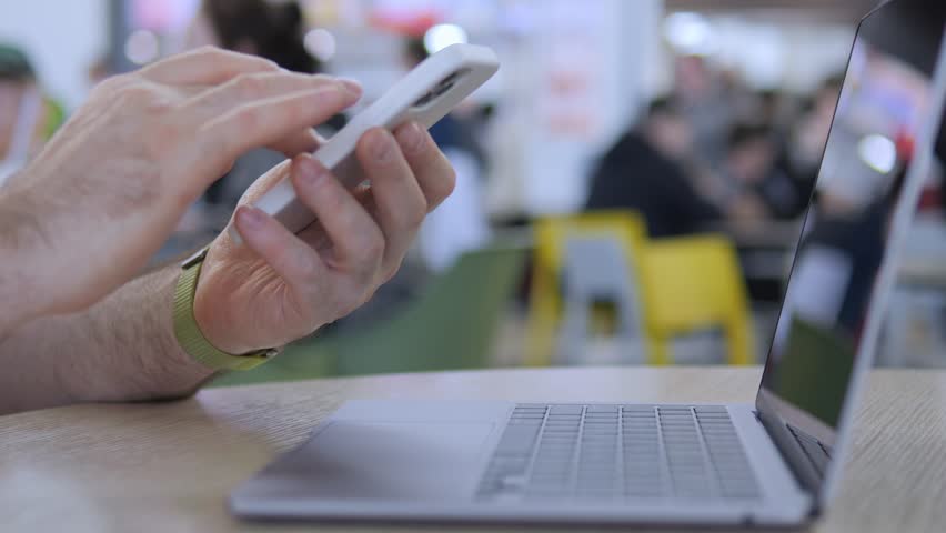 Hands tap phone while laptop sits nearby. Person focused on mobile device. Background shows public space with chairs and people. Suitable for tech, work, or multitasking concepts