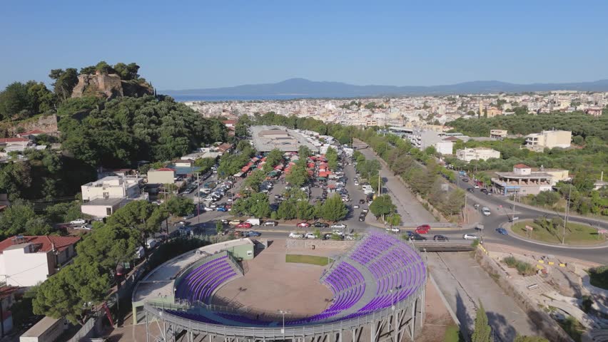 Aerial drone footage of a modern open amphitheater in Kalamata, Peloponnese, Greece, filmed above the market district on a sunny day. The clip shows the amphitheater with distinctive purple seating in the foreground, while the nearby farmer market and covered market complex are visible in the center of the frame. Also visible are a traffic roundabout, surrounding roads, moving cars, parking areas, residential buildings, urban greenery and a broad panoramic view of Kalamata extending toward the mountains and the Mediterranean coastline. Suitable for projects about Greek cities, local markets, public spaces, urban planning, cultural venues, transportation infrastructure, travel and tourism in Southern Europe.