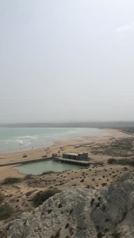 Scenic hilltop view overlooking a sandy beach, ocean waves crashing on rocks, boats, distant houses, and visitors enjoying the shoreline under dramatic cloudy skies and windy weather.