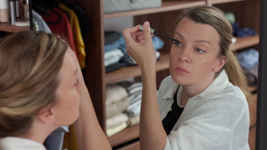 Woman applying mascara while looking in mirror, holding a brush near her eye. Concept of beauty routine, makeup and personal care.