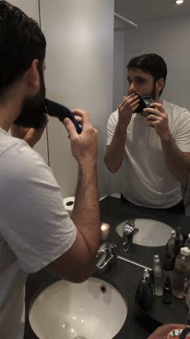 Handsome man in his morning routine trimming and shaping his beard with an electric shaver. Male beauty, personal care and grooming concept in a modern apartment bathroom