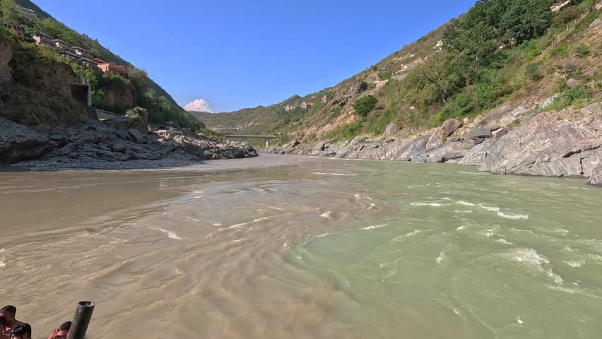 A wide scenic shot showing a dramatic river confluence in a mountainous region. The video features a fast-flowing, sediment-heavy river joining a clearer river alongside a rugged, rocky landscape with distant hillside villages and a bridge.