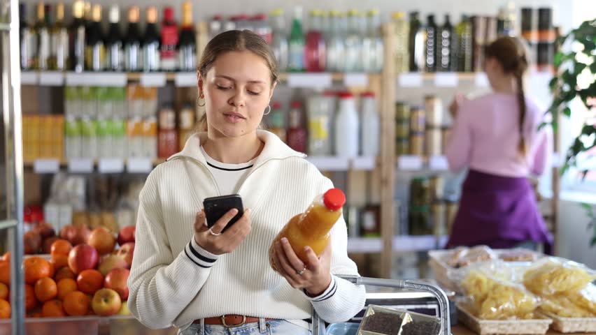 Woman scans the barcode of peach juice on her phone. Customer pays for drinks using a QR code against the background of visitors in a supermarket. High quality 4k footage