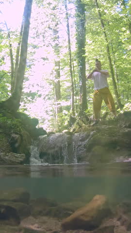 A person balances on a rocky ledge beside a small waterfall in a serene forest, embodying a zen-like pose. The lush greenery and flowing water create a tranquil atmosphise perfect for relaxation.