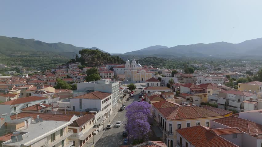 Aerial drone footage of a large Greek Orthodox church in the historic center of Kalamata, Peloponnese, Greece. The scene includes red tiled rooftops, old town streets, Mediterranean urban architecture, a church square, nearby houses and a dramatic mountain landscape in the background. This footage is useful for travel documentaries, tourism promotion, cultural heritage projects, religious architecture content, Greek city visuals, Mediterranean lifestyle videos and urban landscape productions.