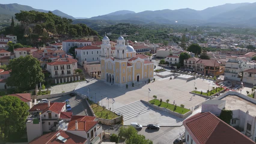 Aerial drone footage of a large Greek Orthodox church with twin bell towers and a spacious church square in the old town area of Kalamata, Peloponnese, Greece. The scene includes red tiled rooftops, historic urban architecture, nearby houses, streets, parked cars, Mediterranean city details and a dramatic mountain landscape in the background. This footage is suitable for travel videos, tourism promotion, cultural heritage projects, religious architecture content, Greek city documentaries, Mediterranean lifestyle visuals and urban landscape productions.