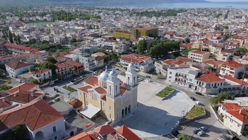 Aerial drone footage of a large Greek Orthodox church with domes and bell towers beside a spacious church square in Kalamata old town, Peloponnese, Greece. The scene includes Mediterranean urban architecture, red tiled rooftops, residential buildings, surrounding streets, parked cars, public space, the sea horizon and mountains in the distance. This footage works well for travel videos, tourism promotion, religious architecture projects, Greek culture documentaries, Mediterranean city visuals, urban heritage content and scenic establishing shots of Kalamata.