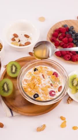 Overhead shot of honey drizzling onto corn flakes, milk, and berries in a glass bowl.