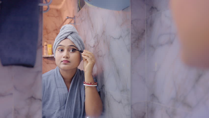 Young lady applying mascara on eyelashes while looking in the mirror, wearing bathrobe in modern bathroom.