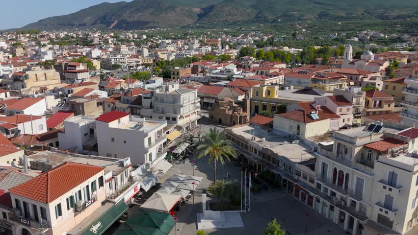 Aerial drone footage approaching an ancient Byzantine church in the old town of Kalamata, Peloponnese, Greece. The scene focuses on the historic stone church surrounded by a public square, traditional buildings, red tiled rooftops, narrow urban streets and local architecture. Green mountains rise behind the city, creating a strong Mediterranean setting for the old town district. This footage is suitable for projects about Byzantine architecture, Greek churches, historic city centers, public squares, Kalamata, travel, tourism, religious landmarks and Southern European urban heritage