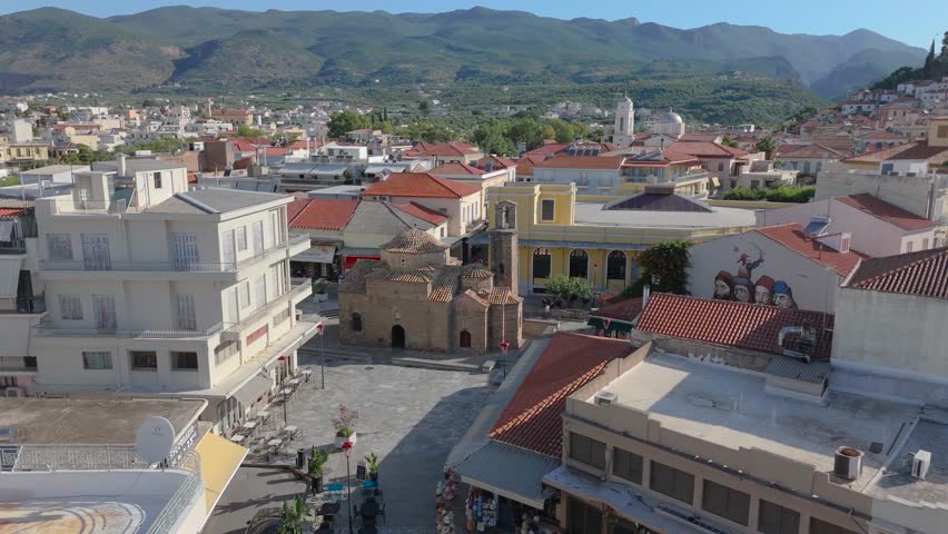 Aerial drone footage of an ancient Byzantine church in the historic center of Kalamata, Peloponnese, Greece. The camera approaches the old stone church surrounded by a paved square, traditional red tiled rooftops, residential buildings, small streets and mountain scenery in the background. The scene captures the atmosphere of Kalamata old town, combining religious heritage, urban architecture, local life and Mediterranean travel. This footage is suitable for projects about Greek landmarks, Byzantine architecture, historic city centers, tourism in Peloponnese, cultural heritage, old towns, traditional architecture and Southern European urban landscapes.