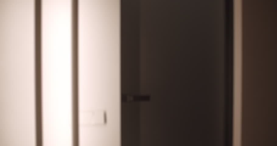 Woman with a ponytail and a tote bag entering a dark apartment at night, her hand touching the wall searching for the light switch before closing the door behind her in slow motion