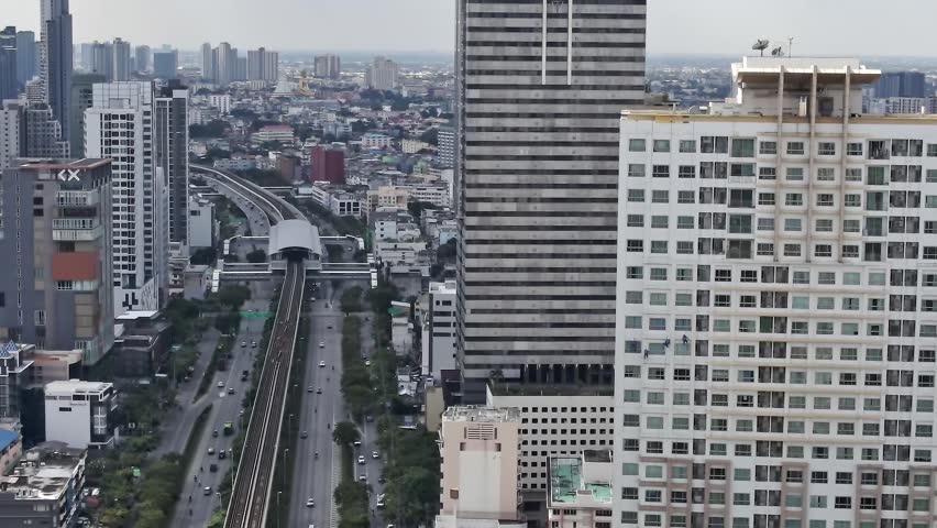 People travel along busy roads in Bangkok. Buildings rise high against a clear sky. The city shows a mix of modern design and urban life. Cars move steadily on the main road beneath.