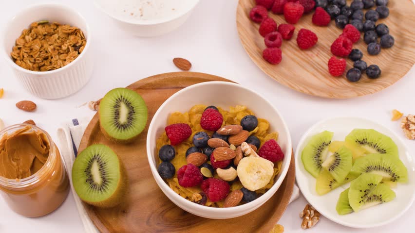 Hand pours crunchy oats into a bowl with berries, kiwi, and nuts on white background.