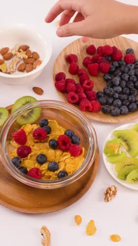 A hand adds nuts to a bowl of cornflakes with berries and kiwi fruit.