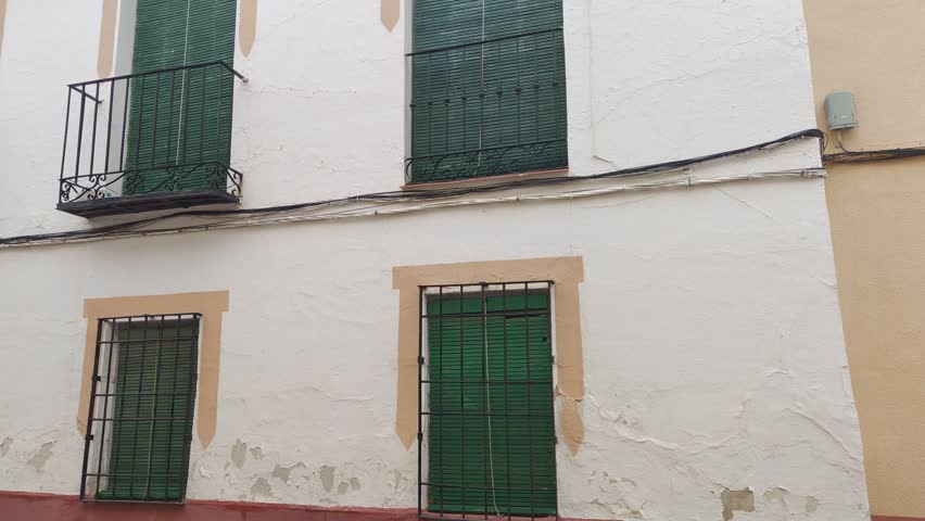 Typical andalusian architecture featuring a white facade with peeling paint, green shutters, and wrought iron balconies on a narrow, traditional cobblestone street in a quiet village in spain